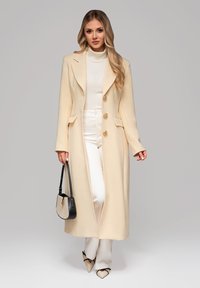 Cappotto lungo crema con tre bottoni dorati, rever a punta e tasche laterali, abbinato a un dolcevita bianco e pantaloni crema. Borsa nera, scarpe a punta.
