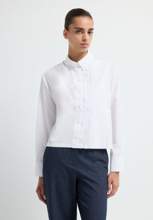 Donna che indossa una camicia bianca con bottoni e tasca frontale, insieme a pantaloni blu scuro a righe, in piedi contro uno sfondo chiaro e semplice.
