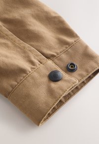 Gros plan sur le poignet d'une chemise en tissu beige avec coutures visibles et un bouton pression noir.