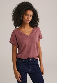 Haut à manches courtes et col en V en bordeaux avec un motif à chevrons texturé, associé à un jean en denim bleu foncé, présenté sur un modèle.