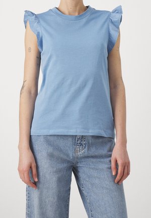Vrouw met een mouwloze lichtblauwe top met ruches op de schouders en lichtblauwe spijkerbroek, staand tegen een effen achtergrond.