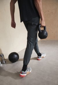 Czarna kettlebell trzymana w jednej ręce, nosząca szare buty sportowe z czerwonymi akcentami, czarne skarpety i dopasowane ciemne spodnie, spacerująca po gładkiej podłodze.