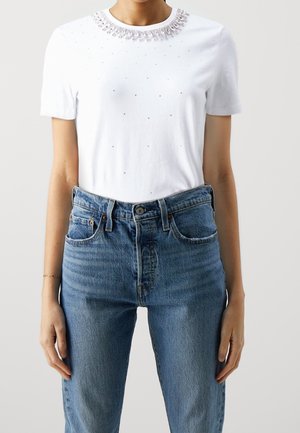 Biała koszulka z krótkim rękawem z koralikowym kołnierzykiem oraz drobnymi, rozrzuconymi koralikami, noszona wpuszczona w niebieskie dżinsy z wysokim stanem.