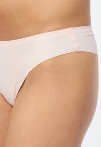 Κοντινό πλάνο στο ισχίο ενός ατόμου που φορά αόρατο ανοιχτό μπεζ εσώρουχο, πάνω σε απλό ανοιχτό φόντο.