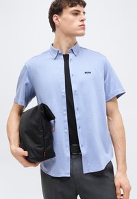 Νεαρός άνδρας φοράει ανοιχτό μπλε πουκάμισο με κοντά μανίκια και το λογότυπο "BOSS", κρατώντας μαύρη τσαντούλα μπροστά σε λευκό φόντο.