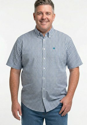 Lächelnder Mann, der ein kurzärmeliges blau-weiß kariertes Hemd und blaue Jeans trägt und mit einer Hand in der Tasche steht.