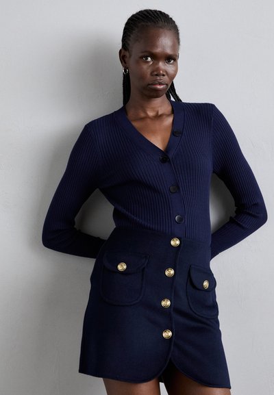 Navy ribbed cardigan with a V-neck and button closure, paired with a navy skirt featuring gold button accents and two front pockets.