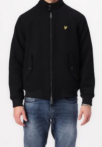 Schwarze Jacke mit Reißverschluss und hohem Kragen, Raglanärmel, zwei vorderen Taschen und einem kleinen gelben Logo auf der linken Brust. Weicher Stoff.