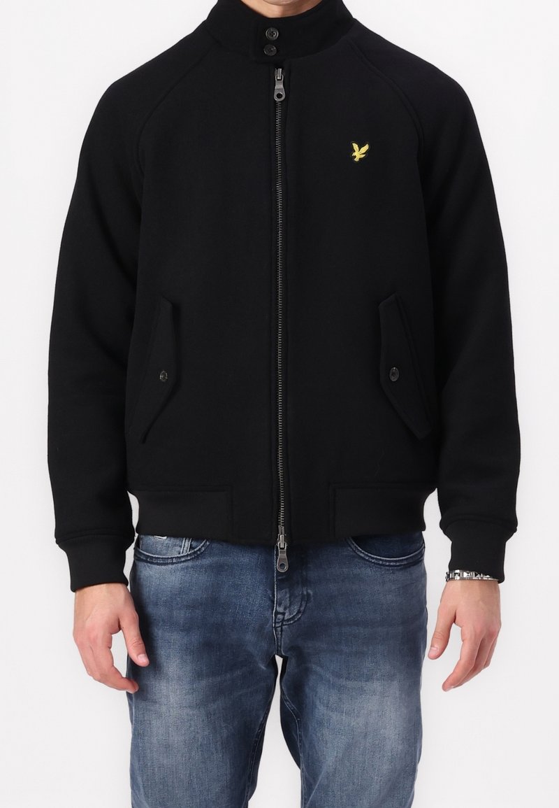 Schwarze Jacke mit Reißverschluss und hohem Kragen, Raglanärmel, zwei vorderen Taschen und einem kleinen gelben Logo auf der linken Brust. Weicher Stoff.
