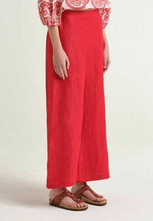 Donna con pantaloni rossi a gamba larga, camicetta rossa e bianca a motivo, e sandali rossi con perline, in piedi davanti a uno sfondo semplice.