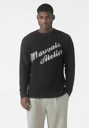 Czarny, miękki sweter dziany z tekstem "Marvvais Atelier" w delikatnym szarym kolorze, z okrągłym dekoltem i długimi rękawami, zestawiony z jasnobeżowymi spodniami.