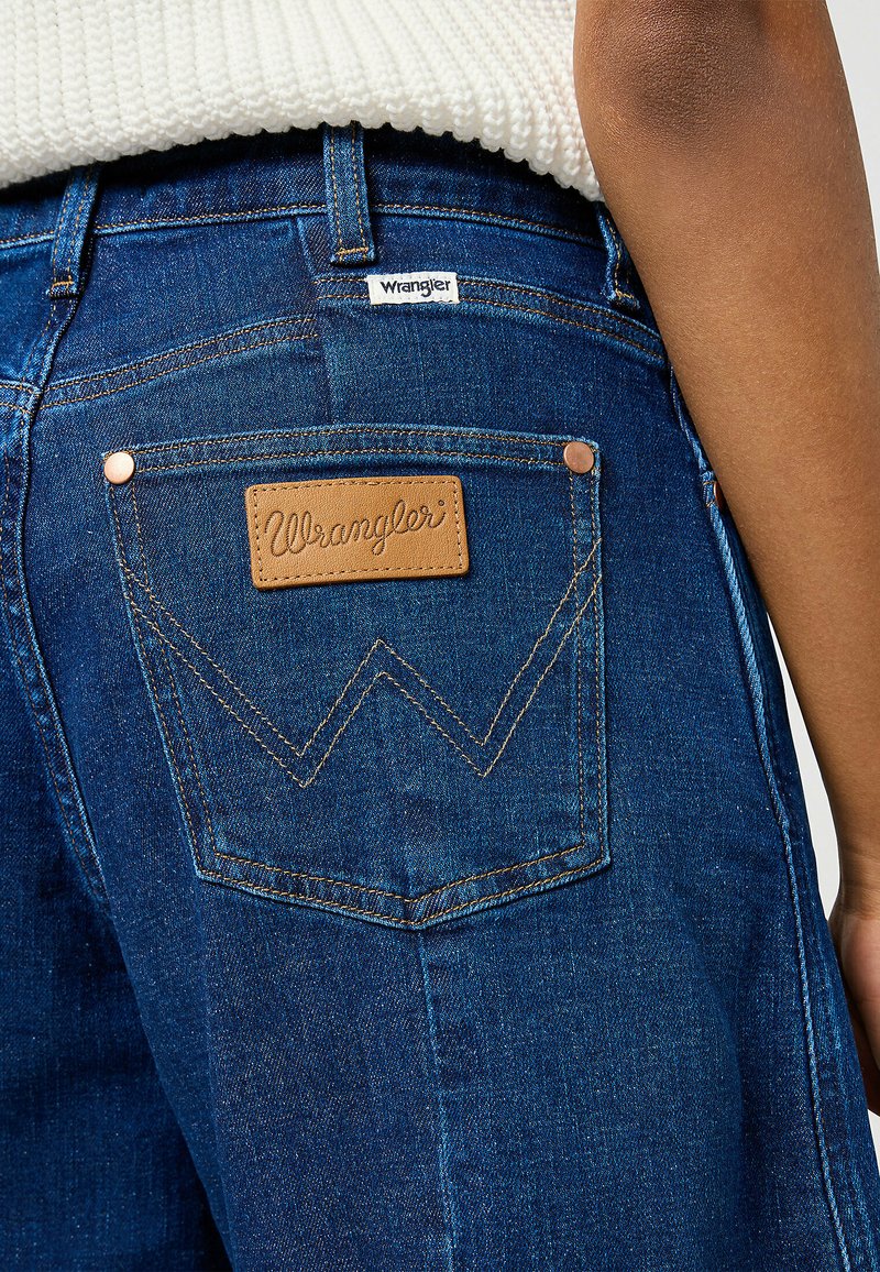 Jean en denim bleu foncé avec un patch en cuir portant "Wrangler", des coutures contrastées et des poches arrière inclinées uniques.