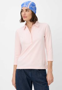 Lyserød polo skjorte med langærmer, krave og knaplukning, parret med mørk denim bukser. Har en glat, bomuldsagtig tekstur.