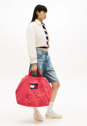 Jeune femme en veste blanche courte, haut à rayures, short en jean et baskets, tenant un grand sac rouge Tommy Jeans par les poignées.