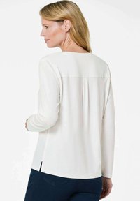 Blusa bianca a maniche lunghe con schiena plissettata, scollatura rotonda, spacchi laterali e texture liscia. Abbinata a jeans blu scuro.