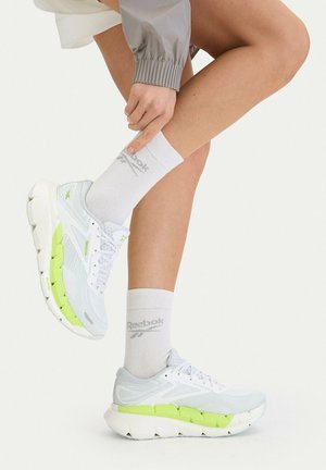 Osoba nosząca białe sneakersy Reebok z neonowozielonymi akcentami oraz białe skarpetki Reebok, dotykająca ręką jednej kostki.