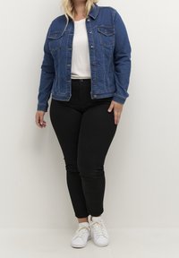 Blå denimjakke med knaplukning, to brystlommer og lange ærmer. Kombineret med sorte, tætsiddende bukser og hvide sneakers.