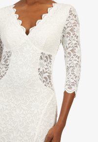 Weißes Spitzenkleid mit gewelltem Ausschnitt, dreiviertellangen Spitzenärmeln und floralem Muster mit transparenten Einsätzen an den Seiten.