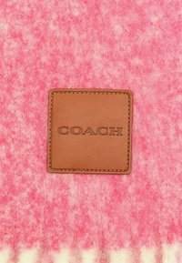 Roze en witte viltstof met een getextureerde bruine leren patch met de opdruk "COACH," die een zachte, fluweelachtige uitstraling benadrukt.