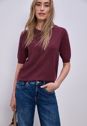 Donna con capelli biondi lunghi che indossa una maglietta a maniche corte color marrone rossiccio e jeans blu, che tiene una borsa in una mano su uno sfondo semplice.