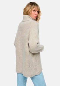 Frau mit blonden Haaren, die einen beigen, grob gestrickten Rollkragenpullover und hellblaue Jeans trägt, leicht zur Seite gedreht mit den Händen in der Nähe der Taille.