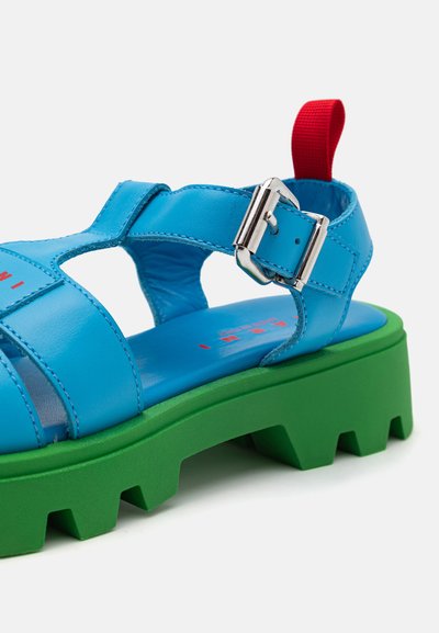 Sandale de piele albastre cu un design deschis, cataramă metalică ajustabilă și talpă chunky verde cu textură pentru aderență. Include o bandă roșie pentru tragere.
