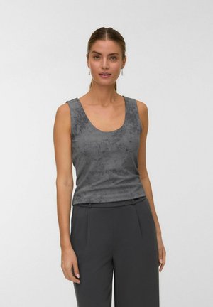 Femme portant un débardeur gris à motifs sans manches et un pantalon taille haute gris foncé, se tenant contre un fond uni.