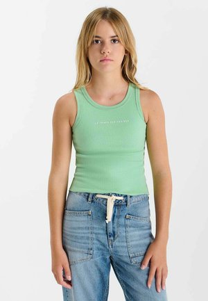 Jeune femme aux cheveux blonds raides portant un haut côtelé vert sans manches et un jean bleu avec des poches avant et une ceinture blanche à cordon.