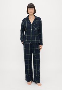 Navy ruudullinen pyjamapuku, joka on valmistettu pehmeästä kankaasta. Pitkähihainen nappipaita taskulla, yhdistettynä löysiin housuihin, joissa on leveät lahkeet.