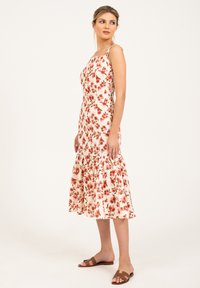 Robe midi florale avec des bretelles fines, fond blanc, motifs de fleurs rouges et vertes, ourlet volanté, associée à des sandales à glissière brunes.
