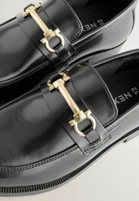 Mocassins en cuir noir présentant un accent en métal doré, avec une finition lisse, des détails cousus, et une semelle texturée.