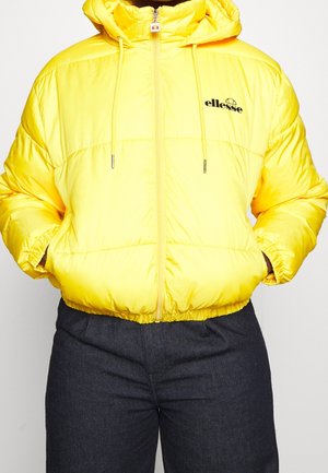 Helle gelbe gesteppte Jacke mit Kapuze, Frontreißverschluss, Kordelzug, Logo auf der Brust, Hände in den Taschen, kombiniert mit dunkler Hose.