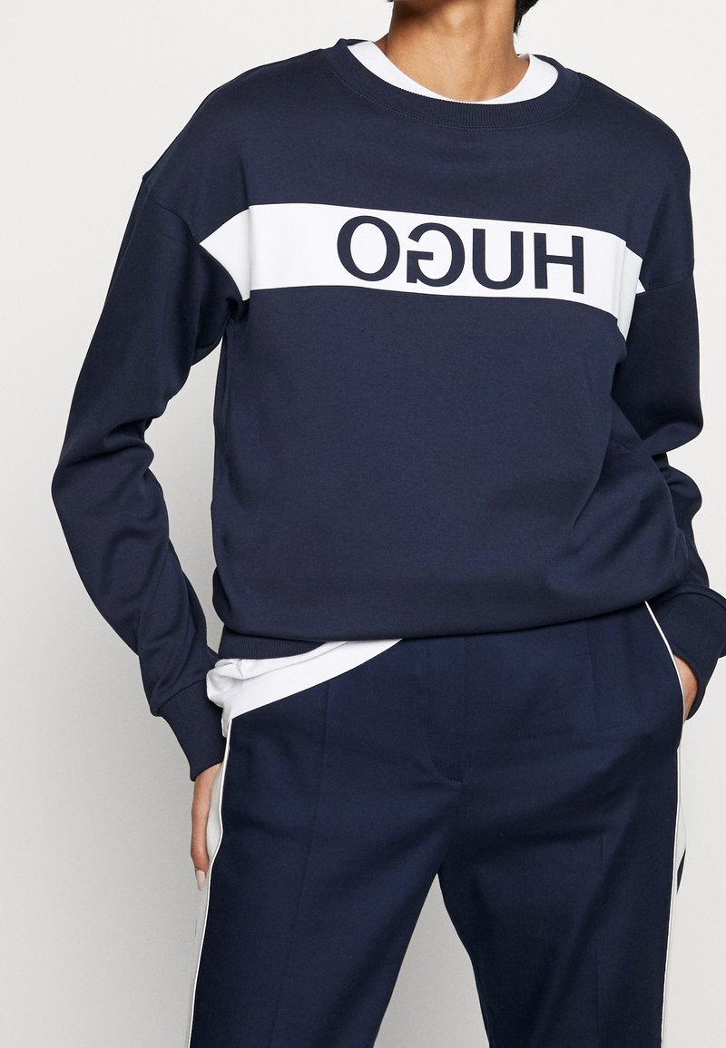 Personne portant un sweat-shirt bleu marine avec une bande blanche et un logo "HUGO" inversé, associé à un pantalon bleu marine assorti avec des bandes blanches sur les côtés.