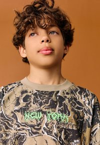 Hanorac cu imprimeu grafic cu craniu și flăcări în bej, negru și verde, cu guler rotund striat. Fundalul este maro solid.