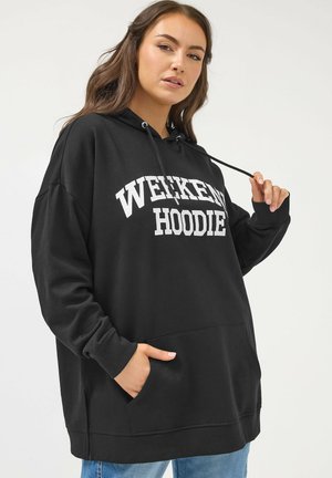 Kobieta w za dużej czarnej bluzie z napisem „WEEKEND HOODIE”, trzymająca jedną sznurek, z ręką w kieszeni na neutralnym tle.