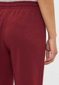 Pantaloni della tuta bordeaux con una texture morbida, vita elastica e cucitura laterale; mostrati da dietro per evidenziare la vestibilità e il design.