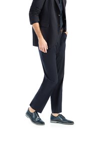 Homme en costume bleu foncé avec pantalon assorti et chaussures habillées à lacets, une main dans la poche, marchant sur fond blanc.