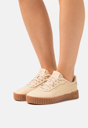 Beige low-top Puma sneakers with gum soles and fuzzy lining worn on feet against a plain background.
