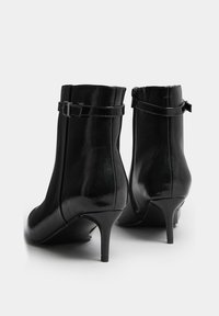 Botines de charol negro con punta afilada, tacón de aguja delgado y detalle de correa ajustable alrededor del tobillo. Cierre de cremallera en la parte posterior.