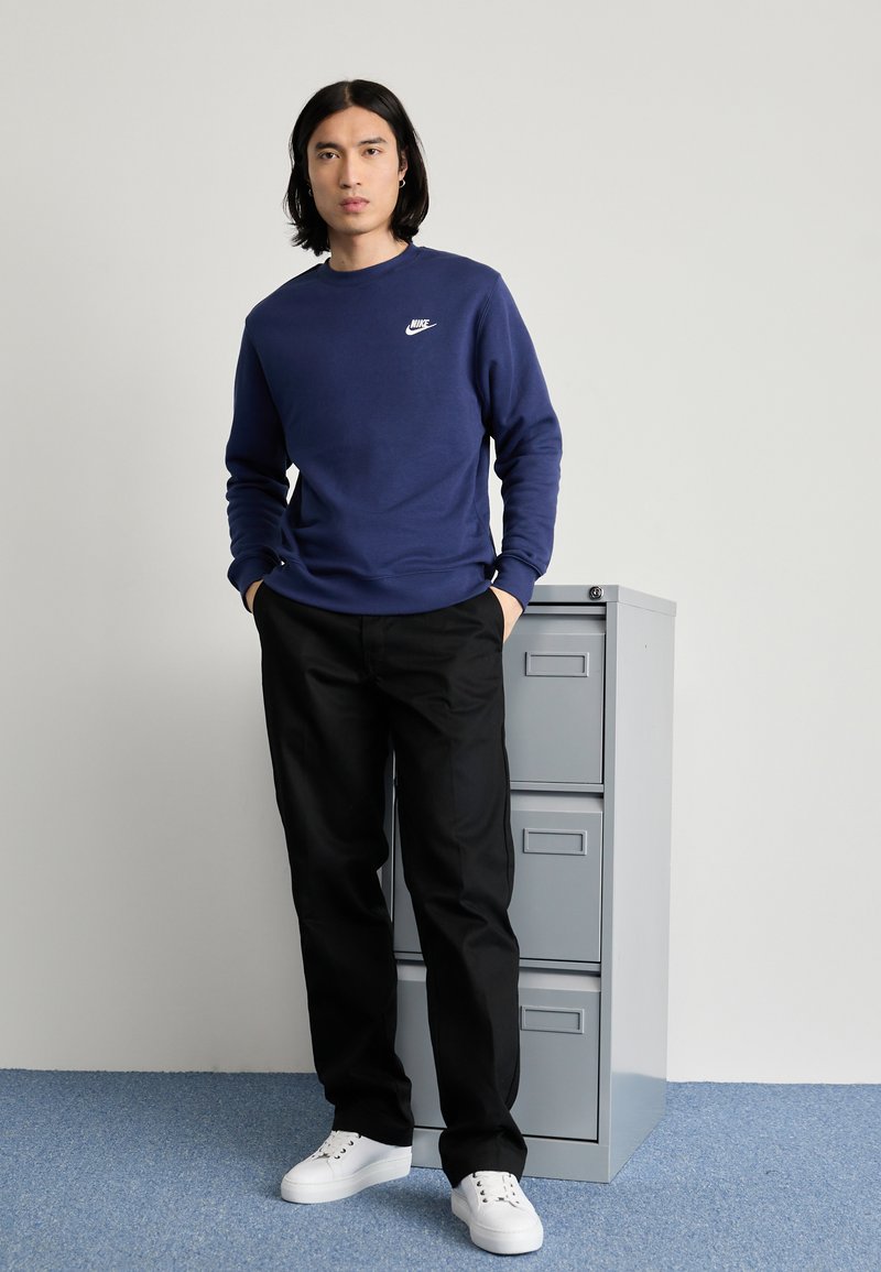 Mörkblå sweatshirt med rund hals och vitt Nike-logotyp, ihopparad med svarta byxor och vita sneakers, stående bredvid en grå Expeditionsskåp.
