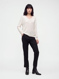 Femme portant un pull crème à col en V et manches longues, pantalon droit noir et bottines noires, debout avec une main dans la poche.