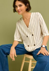 Crème blouse met knoopsluiting, met verticale marineblauwe strepen, korte mouwen en een diepe V-hals, gecombineerd met brede blauwe spijkerbroeken.