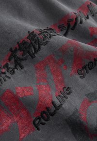 Primo piano di tessuto scuro con testo stampato in rosso e parole ricamate in nero "ROLLING STONE" e caratteri giapponesi.
