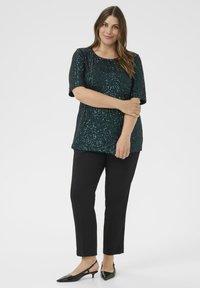Femeie îmbrăcată într-un top scurt cu manșete scurse din paiete teal, pantaloni negri drepți și pantofi cu toc lung și vârf ascuțit negri, stând în fața unui fundal alb.
