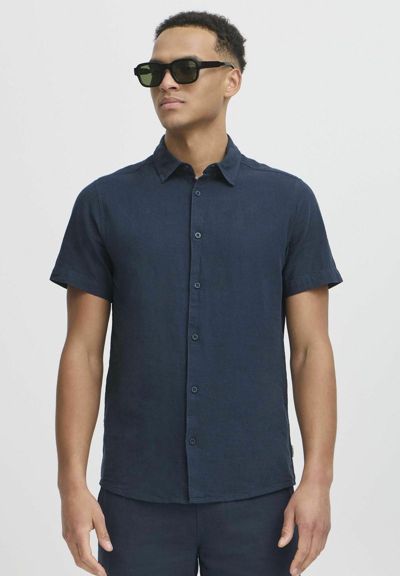 Camisa de lino azul marino de manga corta con botones en el frente, cuello y una textura sutil. Lleva gafas de sol oscuras y está de pie frente a un fondo neutro.