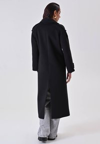 Lungo cappotto nero in tessuto testurizzato, con spacco posteriore, tasche laterali e maniche ampie, abbinato a pantaloni grigi oversize.