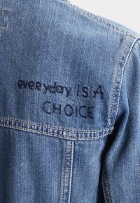 Světle modrá džínová bunda s viditelnými švy a šitím. Na zádech je vyšito heslo „každý den je VOLBA“ tmavě modrou barvou.