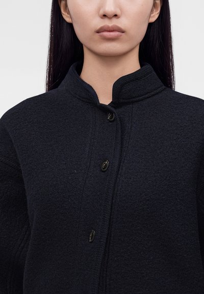 Femme aux longs cheveux foncés portant une veste noire texturée boutonnée avec un col montant sur un fond clair uni.