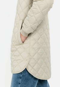 Beige gesteppter Mantel mit einem geometrischen Diamantmuster, seitlichen Taschen und einem abgerundeten Saum. Die Ärmelbündchen haben eine weiche, elastische Textur.