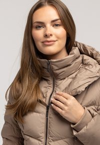 Beżowa kurtka puchowa z wysokim kołnierzem i zapięciem na zamek. Teksturowany materiał, wzór pikowania oraz dzianinowe mankiety. Model trzyma zamek błyskawiczny jedną ręką.